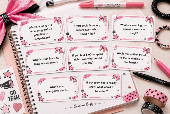 Flat lay image of printable cheer camp conversation starter cards spread across a notebook with pink and black stationery, washi tape, friendship bracelet, and a glitter cheer bow. The printable cards feature fun team bonding questions for cheer camp downtime activities in a modern scrapbook-style aesthetic.