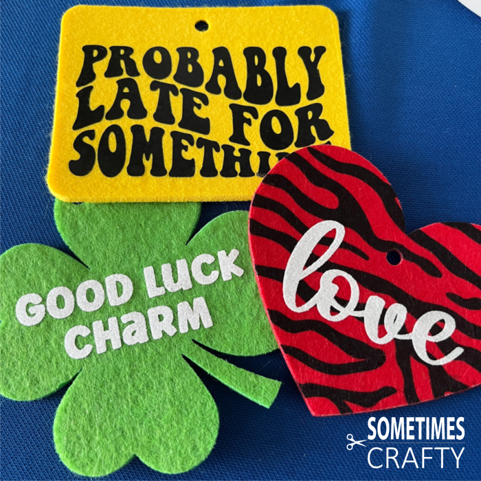 Felt air fresheners in a pile: Horizontal yellow rectangle with black text "Probably late for something", green four leaf clover that says "Good Luck Charm" in white glitter, and a red heart with black tiger stripes that says "love" in a glitter white script font.